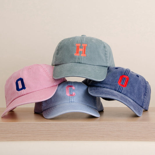 Four kids baseball caps in pink, light blue, medium blue, and navy blue, stacked on a light wood surface against a warm beige background. Each cap features a single embroidered letter logo on the front.