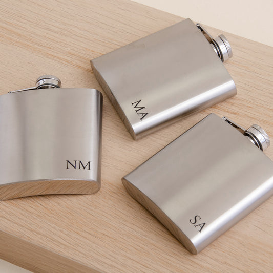 Three brushed silver stainless steel hip flasks with engraved initials (NM, MA, SA) arranged on a light wood grain surface.