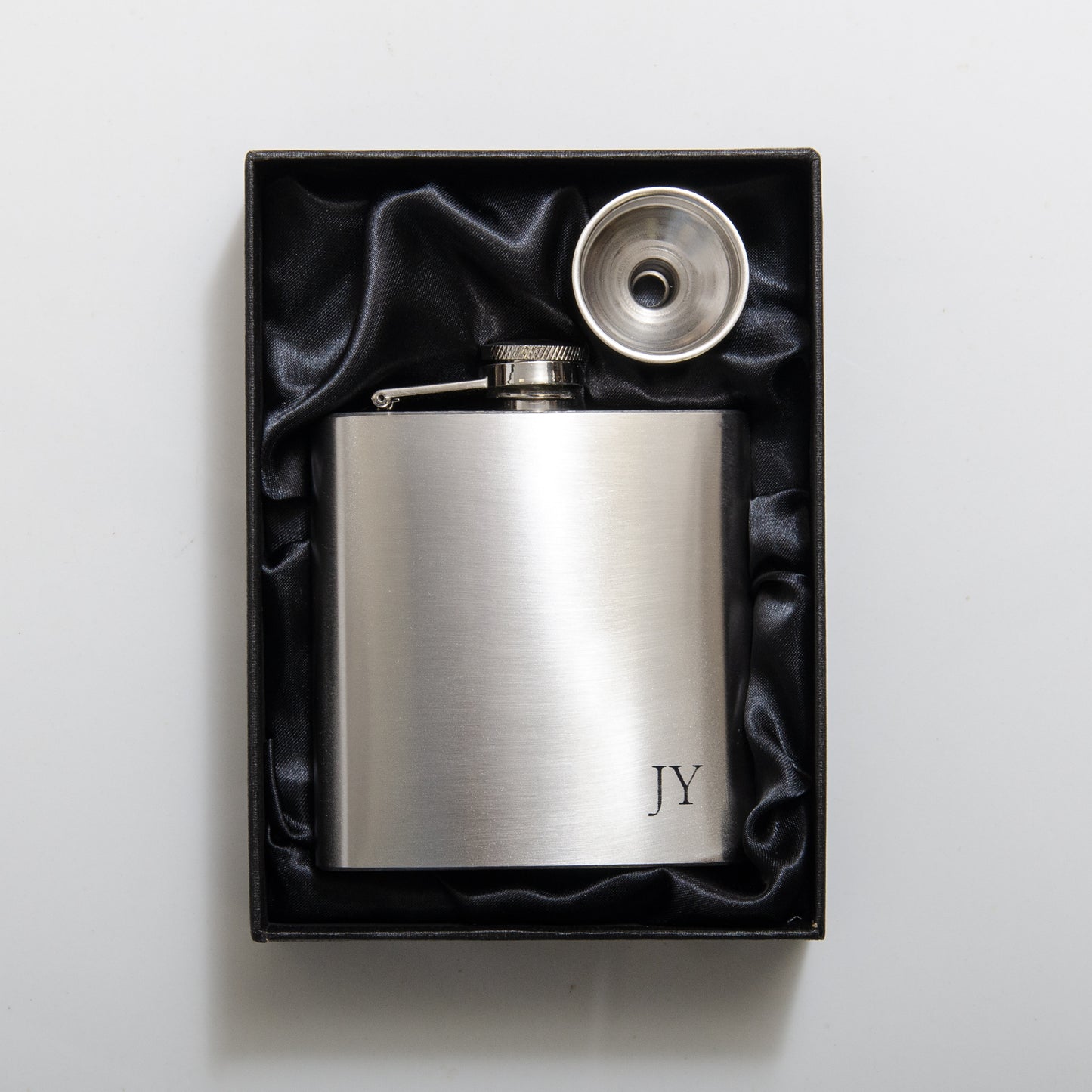 A stainless steel hip flask with engraved initials 'JY' presented in a black box with a stainless steel funnel.