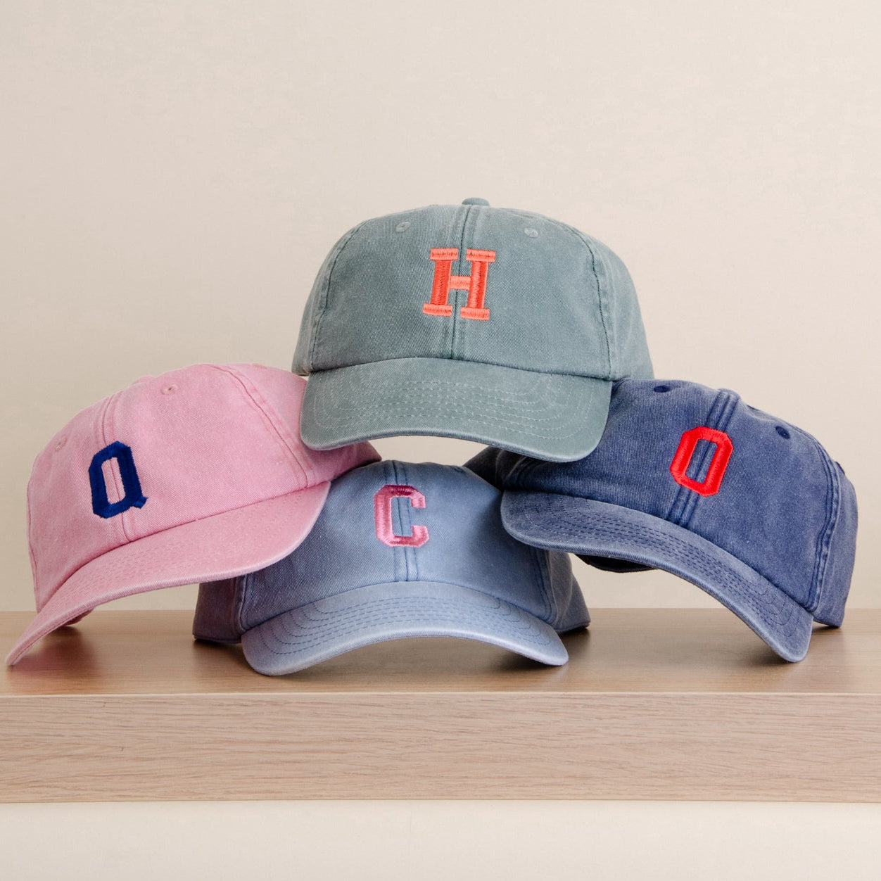 Four kids baseball caps in pink, light blue, medium blue, and navy blue, stacked on a light wood surface against a warm beige background. Each cap features a single embroidered letter logo on the front.