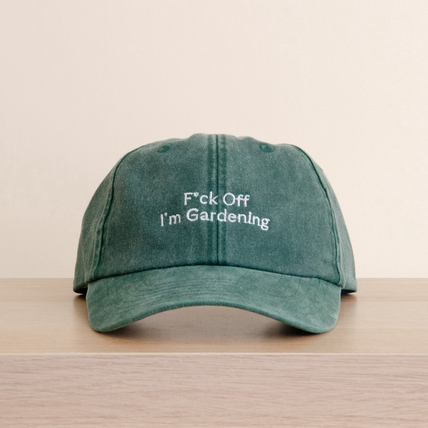 A green washed baseball cap with white embroidered text reading 'F*ck Off I'm Gardenling' resting on a light wood surface against a beige background. Jude Bellingham cap