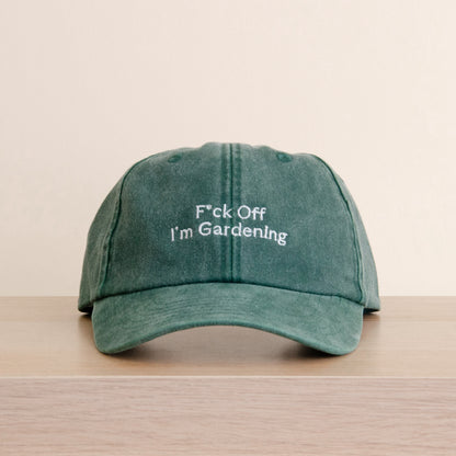 A green washed baseball cap with white embroidered text reading 'F*ck Off I'm Gardenling' resting on a light wood surface against a beige background. Jude Bellingham cap