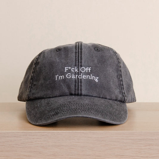 A dark gray washed baseball cap with white embroidered text reading 'F*ck Off I'm Gardenling' resting on a light wood surface against a beige background. Jude Bellingham cap