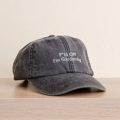 A black washed baseball cap with white embroidered text reading 'F*ck Off I'm Gardening', resting on a light wood surface against a plain beige background.
