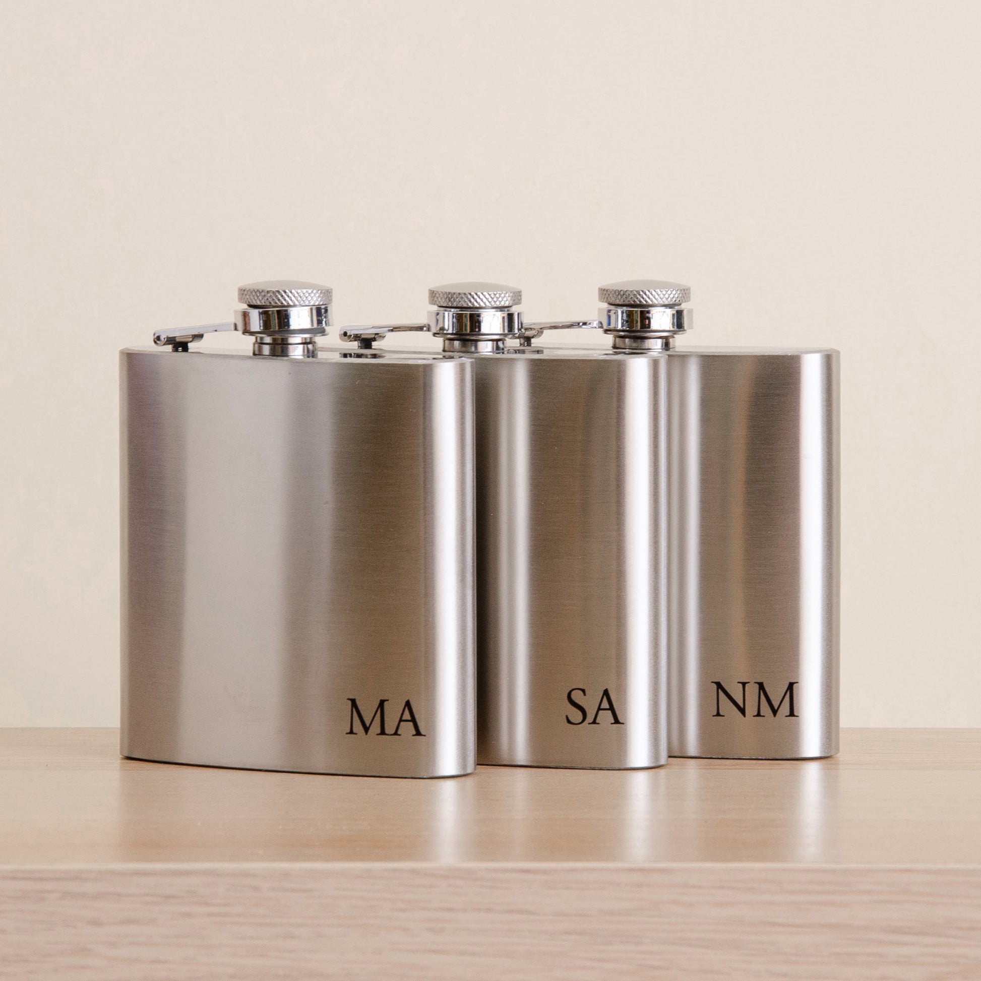Three stainless steel hip flasks with silver screw-top caps, arranged side by side on a light wood surface against a warm beige background. The flasks are engraved with the initials MA, SA, and NM.