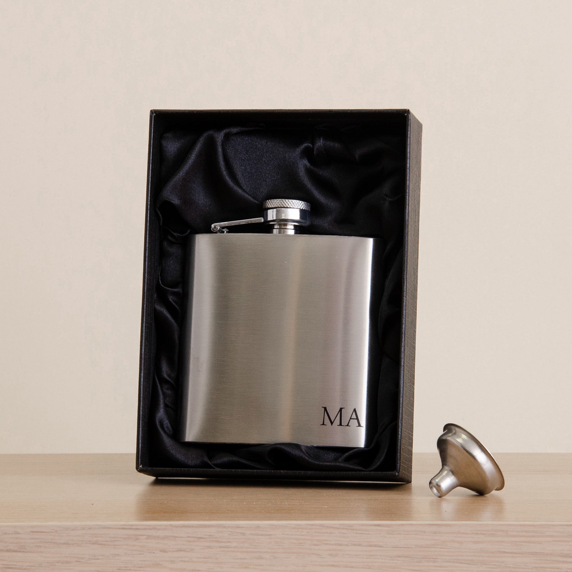 A brushed stainless steel flask with a silver cap, personalized with the initials 'MA' in black, presented in a black gift box with black satin lining. The flask sits on a light wood surface against a warm beige background, with the cap resting beside the box.
