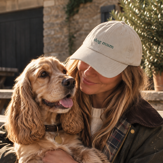 A woman outdoors holds a brown Cocker Spaniel dog close to her chest. She is wearing a beige baseball cap with green embroidered text that reads 'dog mum' and a dark olive jacket.