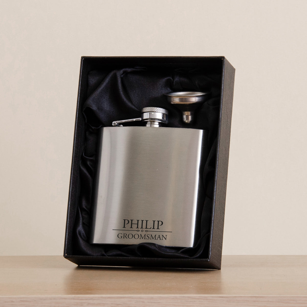 A personalized stainless steel flask engraved with 'PHILIP GROOMSMAN', presented inside an open black gift box with black satin lining on a light wood surface against a beige background.