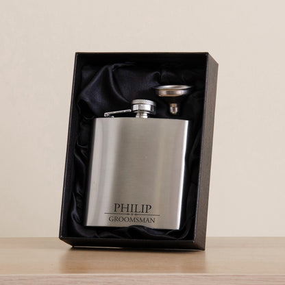 A personalized stainless steel flask engraved with 'PHILIP GROOMSMAN', presented inside an open black gift box with black satin lining on a light wood surface against a beige background.