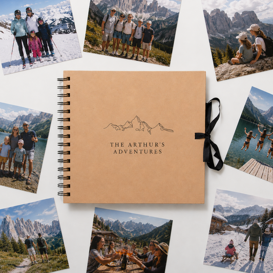 A spiral-bound photo album with a tan cover lies on a white surface, surrounded by scattered travel photographs. The center cover shows a simple mountain line drawing and the text 'THE ARTHUR'S ADVENTURES.