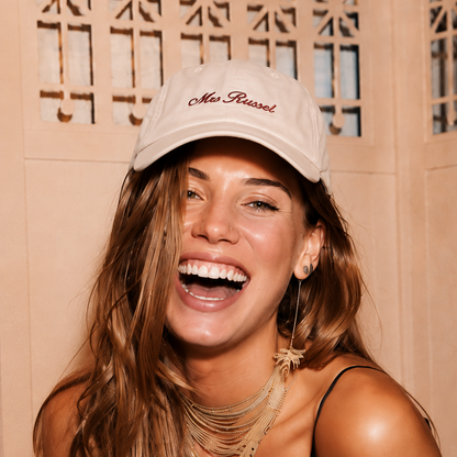 A woman with long light brown hair smiling broadly, wearing a beige baseball cap with 'Mia Russell' embroidered in red script, a black spaghetti-strap top, layered gold necklaces, and a white fringed cardigan. She is posed in front of a beige wooden screen background.
