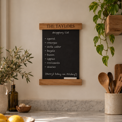 A black chalkboard shopping list mounted on a beige wall in a warm, modern kitchen. The chalkboard has a light wood top and bottom frame with the text 'THE TAYLORS' at the top.