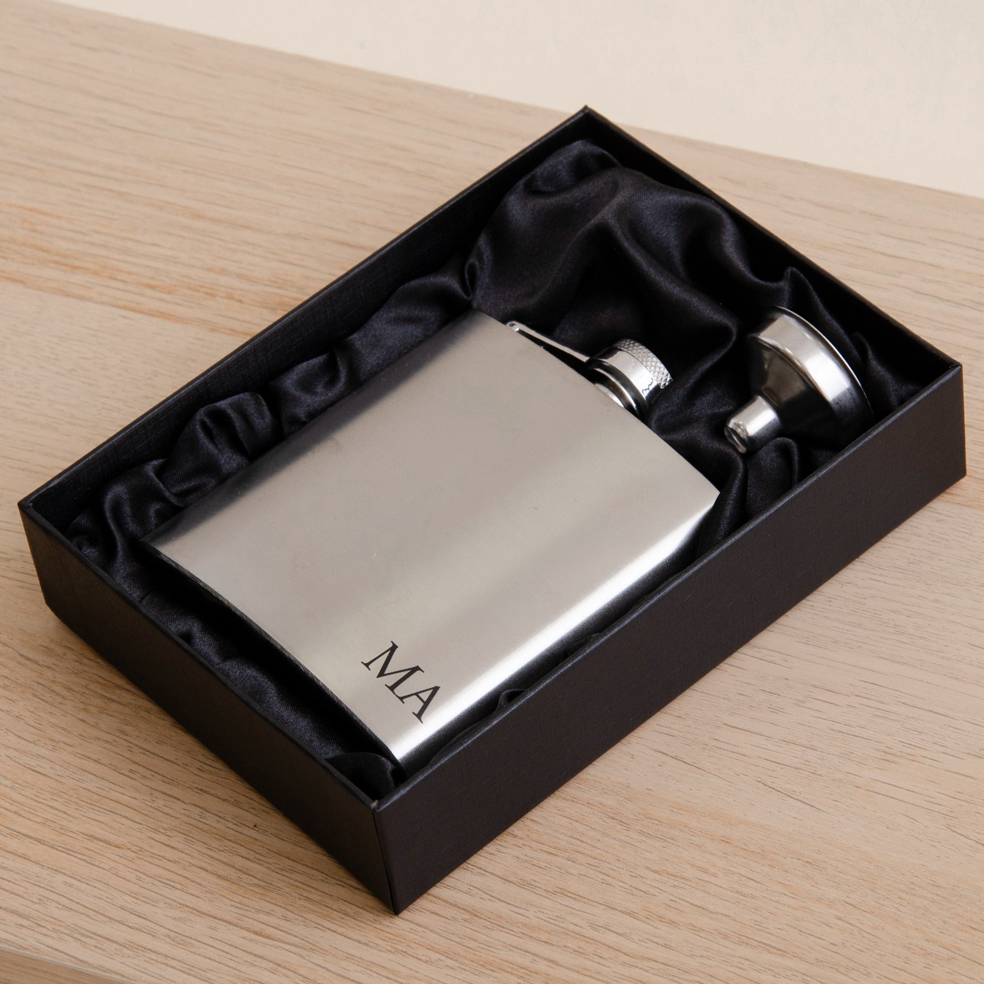 A polished silver stainless steel flask with 'MA' engraved on the front, presented in a black gift box with black satin lining on a light wood tabletop.