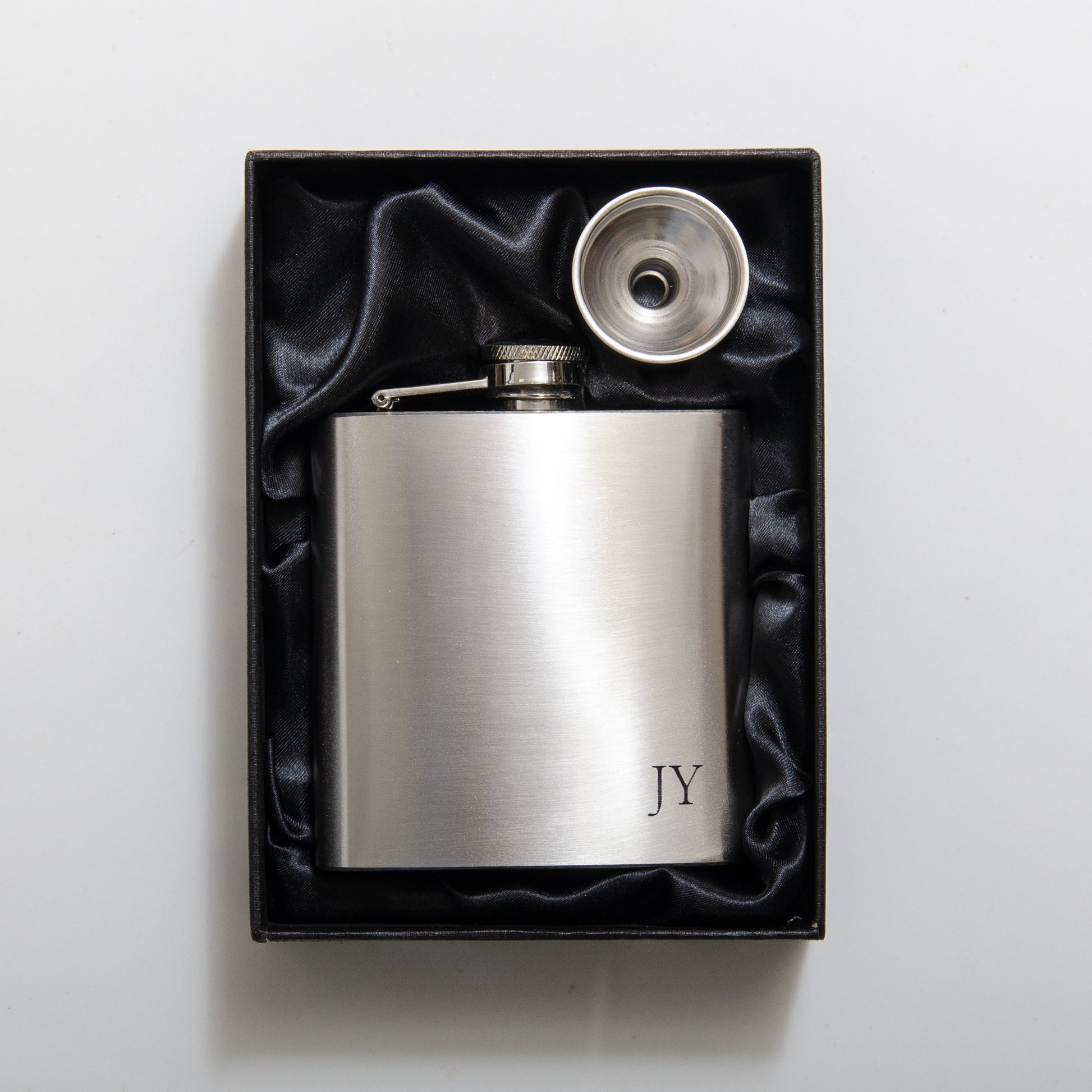 A stainless steel hip flask with engraved initials 'JY' presented in a black box with a stainless steel funnel.