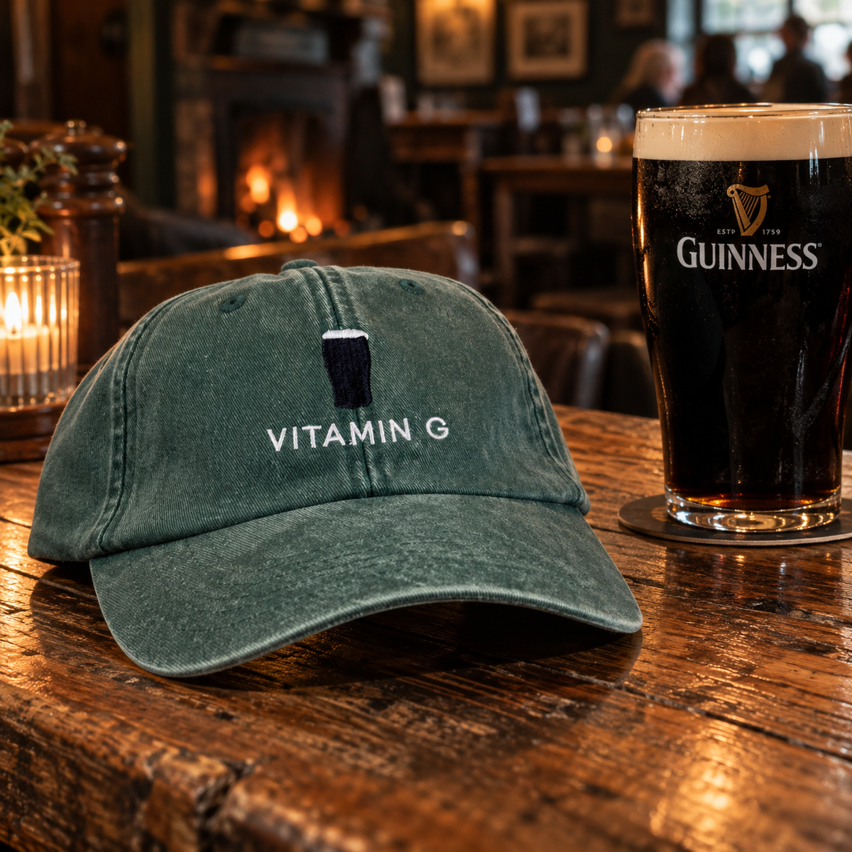 A dark green washed baseball cap with 'VITAMIN G' and a pint glass logo embroidered on the front, resting on a rustic wooden bar next to a pint of Guinness in a cozy pub setting.