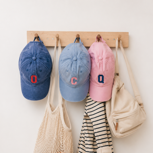 Three baseball caps in blue, light blue, and pink hang on a wooden rack with cream-colored bags and a striped garment beside them against a plain white wall.